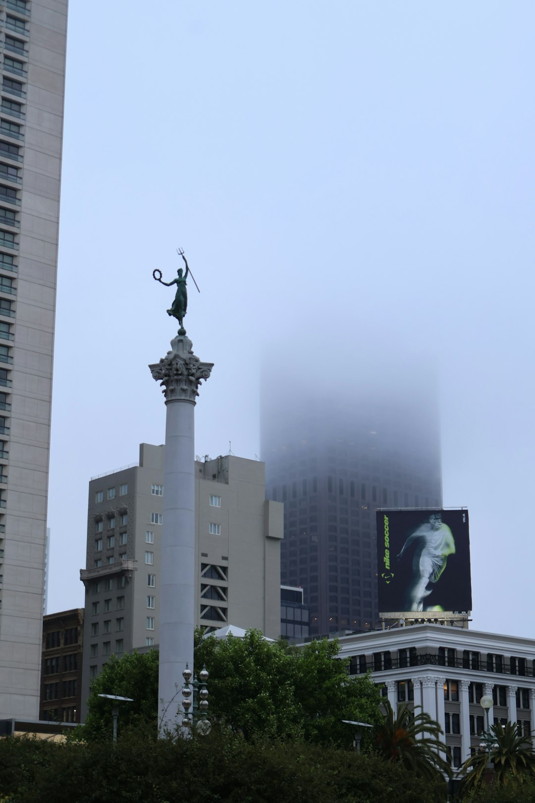Fog-shrouded Dewey Monument statue at Union Square in San Francisco, California, with the figure atop a tall column rising above skyscrapers, palm trees, and billboards on a misty day.